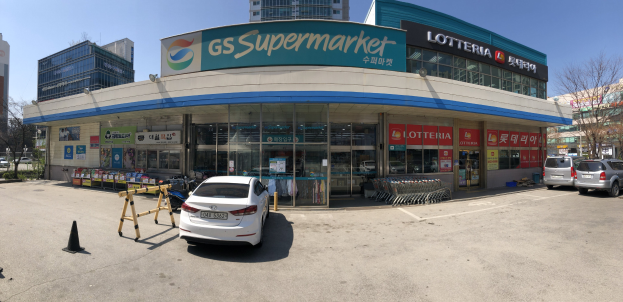 Ein Auto vor einem Supermarkt mit der Aufschrift "GS Supermarkt" geparkt, umgeben von anderen Fahrzeugen, Verkehrskegeln und verstreuten Gegenständen auf der Straße, vor einer Kulisse aus Gebäuden, Bäumen und einem klaren blauen Himmel.