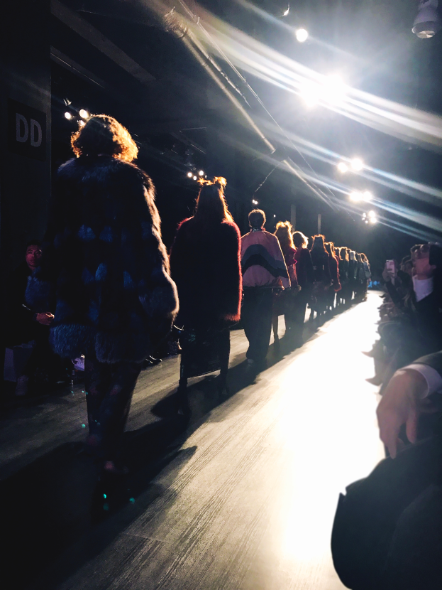 Eine Gruppe von Menschen läuft über den Laufsteg einer Modenschau, beleuchtet von hellen Scheinwerfern, mit Zuschauern auf beiden Seiten.