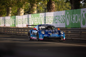Blauer Porsche 911 GTS auf einer Rennstrecke mit einem Zaun und Bannern im Hintergrund, Bäume und einen klaren Himmel.