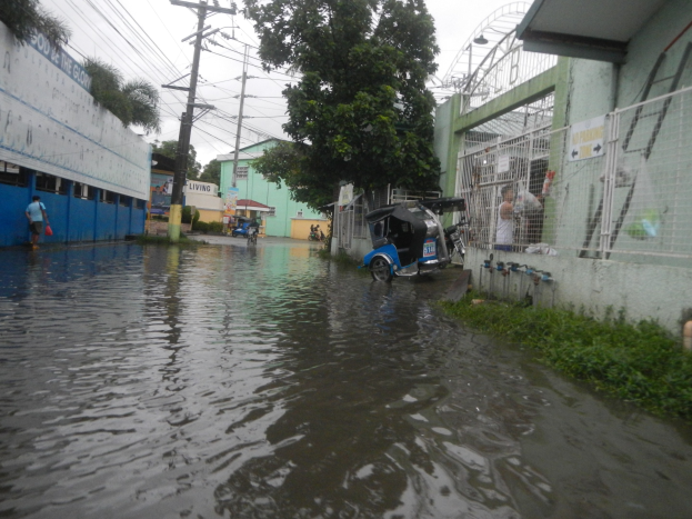 Eine überflutete Straße mit einem geparkten Motorrad in der Mitte, Gebäude, Bäume und Strommasten auf beiden Seiten sowie einige Menschen und Fahrzeuge im Hintergrund.
