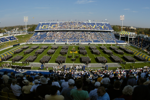 Eine große Menschenmenge in einem Stadion bei einem Fußballspiel, einige stehen, andere sitzen, auf einem Rasenfeld unter einem klaren blauen Himmel, mit Laternenmasten, Bäumen und Gebäuden im Hintergrund.