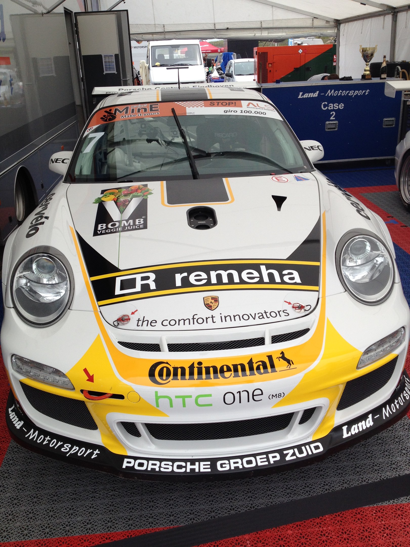 Weißer und gelber Porsche 911 GT3 RSR Rennwagen in einer Garage mit anderen Fahrzeugen, Menschen und Gegenständen geparkt.