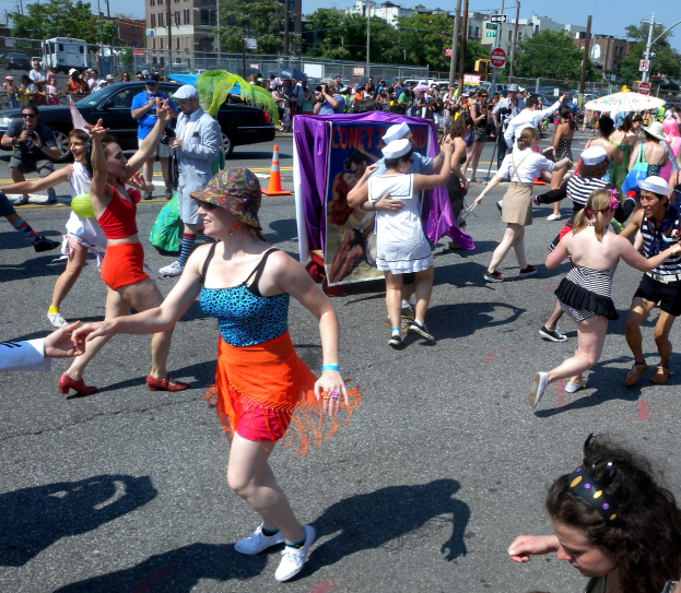Eine Gruppe von Menschen, die eine Stra√e entlangmarschiert, einige mit Mötzen und Schirmen, andere tanzen, mit einem Banner, Verkehrskegeln, Fahrzeugen, Lichtmästen, Schildern, Bäumen, Gebäuden und einem klaren blauen Himmel im Hintergrund.
