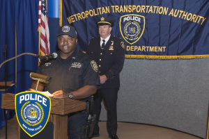 Zwei Polizeibeamte auf einem Podium mit einer Fahne, einem Banner und einer Tafel mit Symbolen und Text, einer Beamter spricht in ein Mikrofon und hält Papers.