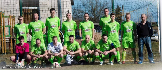 Eine Gruppe von Männern in grünen T-Shirts steht und hockt auf einem Fußballfeld mit einem Ball davor, ein Torpfosten rechts und ein Hintergrund aus Bäumen, Gebäuden, Pfählen, Fahrzeugen und dem Himmel, mit einer Zahlenfolge links unten.