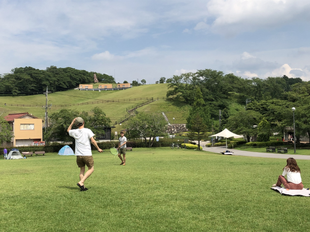 Gruppe von Menschen, die Badminton in einem Park spielt, mit einem Mann, der einen Schläger hält und auf einer Decke auf dem Gras sitzt, in der Nähe von Zelten und Gebäuden mit Hügeln und bewölktem Himmel im Hintergrund.