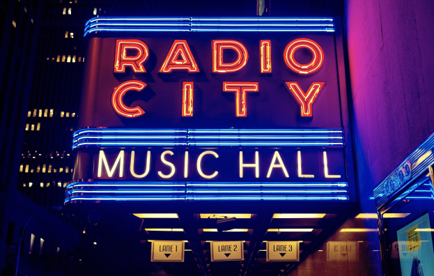 Radio City Music Hall in New York City, hell erleuchtet in der Nacht, umgeben von hohen Gebäuden, mit einer 'Radio City'-Leuchtschrift und zusätzlichen Schildern auf der rechten Seite.