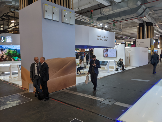 Eine Gruppe von Menschen geht um den Messestand der UAE Space Agency herum, mit Taschen, Informationsplakaten, Bildschirmen und anderen Gegenständen im Hintergrund, unter Deckenleuchten und Säulen.
