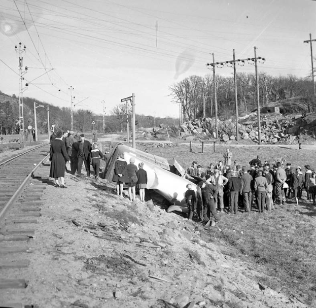 Eine Gruppe von Menschen steht um einen Zug herum, der auf einem Bahngleis in einen Felsenkranz gekracht ist. Im Hintergrund sind Strommasten und -leitungen zu sehen, sowie Bäume und Himmel in der Ferne. Das Bild ist in Schwarz-Weiß.