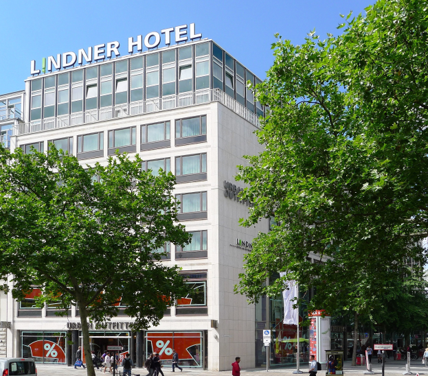 Großes modernes Hotelgebäude mit Glasfenstern und Beschriftung oben, umgeben von Bäumen und einer belebten Straße mit Fußgängern und Fahrzeugen in Berlin, Deutschland.