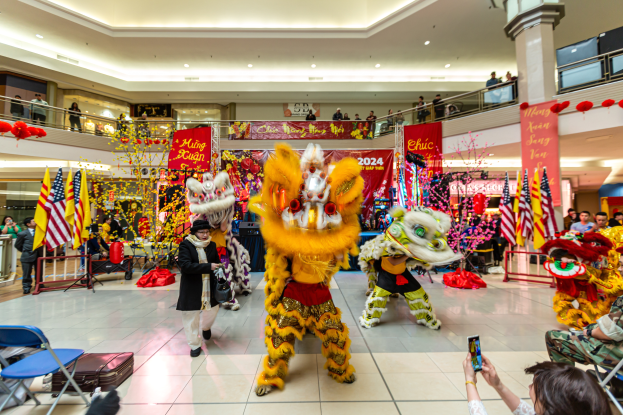 Lebendiges chinesisches Neujahrsfest in einem Einkaufszentrum mit Menschen in Kostümen, die tanzen, und anderen, die auf Stühlen sitzen und Mobiltelefone halten, umgeben von Fahnen, Bannern, Dekorationen und Lichtern.