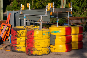 Ein Haufen gelber und roter Reifen auf einem Schmutzfeld umgeben von verstreuten Metallobjekten, Brettern und anderem Schutt, mit Bäumen, Pflanzen und einem Schuppen im Hintergrund.