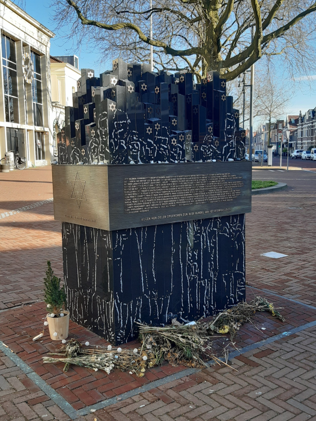 Denkmal in einem Stadtplatz für Holocaust-Opfer, mit einem Baum davor, einem Topfpflanze auf dem Boden, verstreuten trockenen Blättern und Gebäuden, Fahrzeugen, Pfählen und Bäumen im Hintergrund.