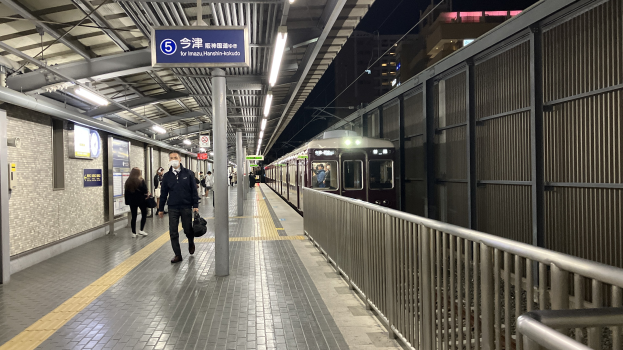 Ein Zug fährt auf ein Bahnsteiggelände zu, mit Menschen in der Nähe, einem Geländer auf der rechten Seite, Gebäuden im Hintergrund, Bahnsteigbeleuchtung und einer Abfahrtsanzeige.