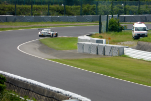 Ein Rennwagen fährt auf einer Rennstrecke mit einem Polizeiwagen im Hintergrund, umgeben von Gras, Pflanzen, Bäumen und Absperrungen, mit einem Fahrzeug auf der rechten Seite und Text am unteren Rand.