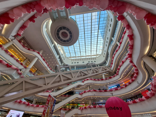 Ein Einkaufszentrum im Valentinstag-Dekor mit pinken und weißen Luftballons, Rolltreppen, Geländern, Texttafeln, Lichtern und einer Glasdecke.