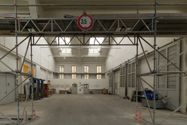 Lagerhalle mit ausführlichem Gerüst, Verkehrskegel, Metallstangen, ein Schild, Maschinen, eine Tür, Fenster, Wände, Deckenleuchten und Dach.