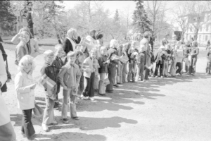 Schwarz-weißes Foto von Menschen in einer Reihe auf einer Schotterstraße, die Fahnen schwenken, während sie an einer Schule an einem Protestmarsch teilnehmen, mit Bäumen, Gebäuden und einem klaren Himmel im Hintergrund.