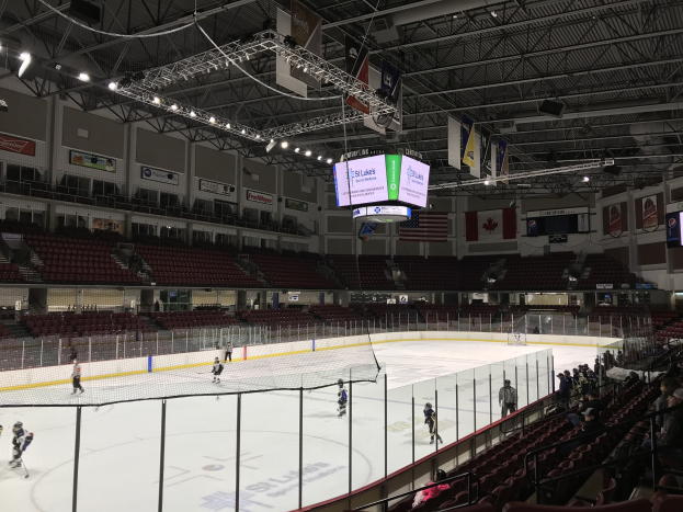 Eishockeyspiel in einer großen Arena mit einem zentralen Netz, Zuschauern auf beiden Seiten, Deckenbeleuchtung und Trägern sowie Bannern an den Wänden, identifiziert als die St. Louis Blues Arena.