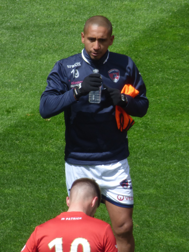 Ein Mann in einem Sportdress steht auf einem grünen Feld und hält eine Flasche in einer Hand und ein oranges Tuch in der anderen.