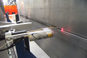 Helle Laserschneidemaschine, die Licht in einer industriellen Umgebung mit einer Wand auf der rechten Seite und Hintergrundobjekten aussendet.