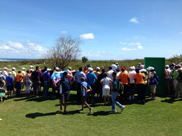 Gruppe von Menschen auf einem grünen Feld mit Bäumen, Pflanzen und einem Gewässer im Hintergrund, wahrscheinlich bei einem Golfturnier.