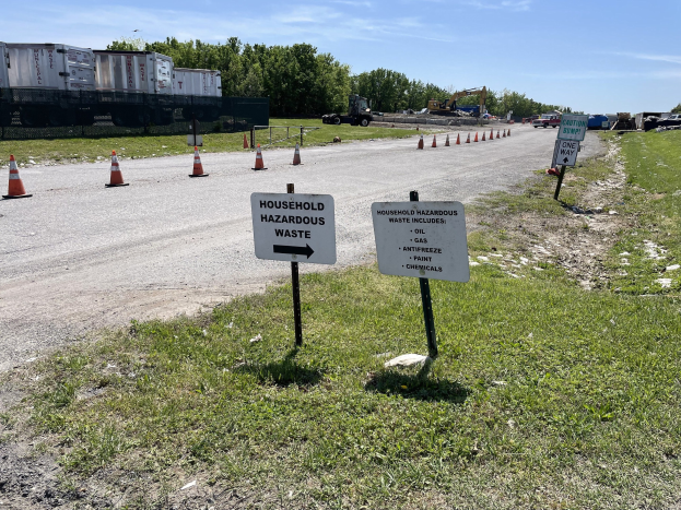 Straßensammelstelle für "Gefährliche Haushaltsreste" markiert durch Verkehrskegel, mit Fahrzeugen, Gras, Bäumen und bewölktem Himmel im Hintergrund.