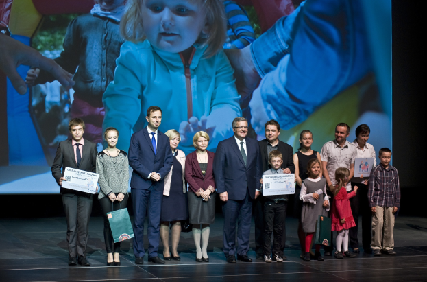 Gruppe von Menschen auf einer Buehne mit Taschen und Urkunden, im Hintergrund ein Bildschirm mit einem Kind und anderen