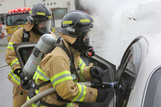 Zwei Feuerwehrleute in Schutzausrüstung verwenden einen Schlauch, um ein brennendes Auto zu löschen, mit mehreren Fahrzeugen und einem Gebäude im Hintergrund, Rauch steigt von dem Auto auf.