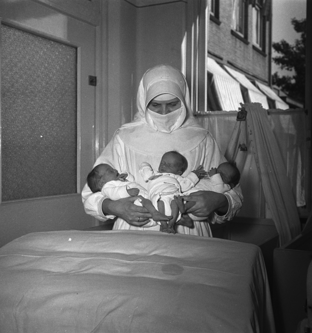 Ein Schwarz-Weiß-Bild einer Frau in einem Krankenhausbett, die zwei Babys hält, mit einer Wand, einer Tür, einem Fenster, einem Baum und einem Gebäude im Hintergrund.