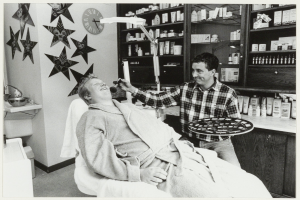 Ein Mann lächelt in einem Zahnarztstuhl, während ein anderer Mann neben ihm steht und ein Tablett hält, mit einem Waschbecken, Sternen, einer Uhr und Regalen mit Flaschen im Hintergrund.