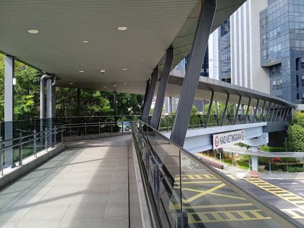 Fussg"angerweg mit Glasgel"ander, beleuchtet von Deckenl"ampen, mit einem Schild, umgeben von Geb"uden, B"umen, Pflanzen, Gras, einer Stra{\ss}e und einer Br"cke im Hintergrund.