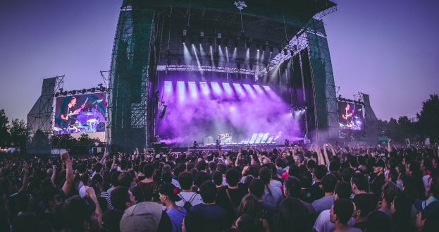 Eine große Menschenmenge versammelte sich auf einem Musikfestival, mit einer Bühne in der Mitte, auf der Musiker Instrumente spielten, beleuchtet von Scheinwerfern und umgeben von Bildschirmen, vor einer Kulisse aus einem Gebäude, Bäumen und einem klaren blauen Himmel.