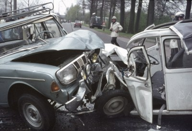 Ein verunglücktes Auto am Straßenrand mit Zuschauern in Helmen und anderen Fahrzeugen im Hintergrund, vor einem Wald, Masten und einem klaren blauen Himmel.