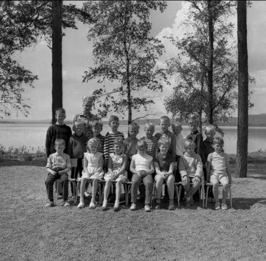 Schwarzes und weißes Foto von Kindern, die auf Stühlen sitzen und vor einem See stehen, mit Bäumen, Pflanzen, Hügeln und einem bewölkten Himmel im Hintergrund, beschriftet mit "1940er-Kinder am See."