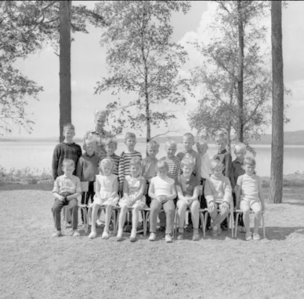 Schwarzes und weißes Foto von Kindern, die auf Stühlen sitzen und vor einem See stehen, mit Bäumen, Pflanzen, Hügeln und einem bewölkten Himmel im Hintergrund, beschriftet mit "1940er-Kinder am See."