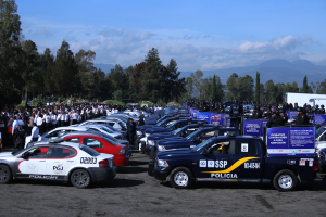Eine große Anzahl von Polizeiwagen auf einem Parkplatz mit Menschen drumherum, Schilder mit Text und eine Kulisse aus Bäumen, Hügeln und einem bewölkten Himmel.