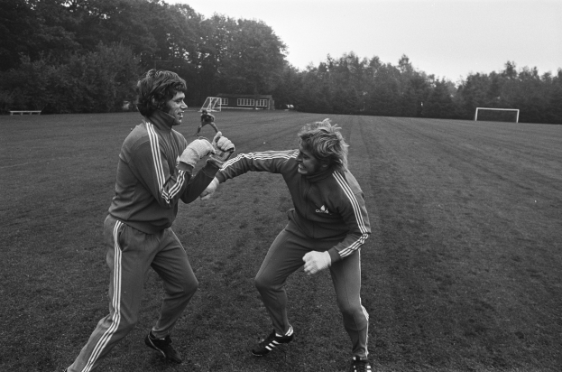 Zwei Männer in Schwarz-Weiß-Uniformen sind in eine körperliche Auseinandersetzung auf einem Fußballfeld verwickelt, umgeben von Bäumen und einer Hütte im Hintergrund, beide tragen Handschuhe.