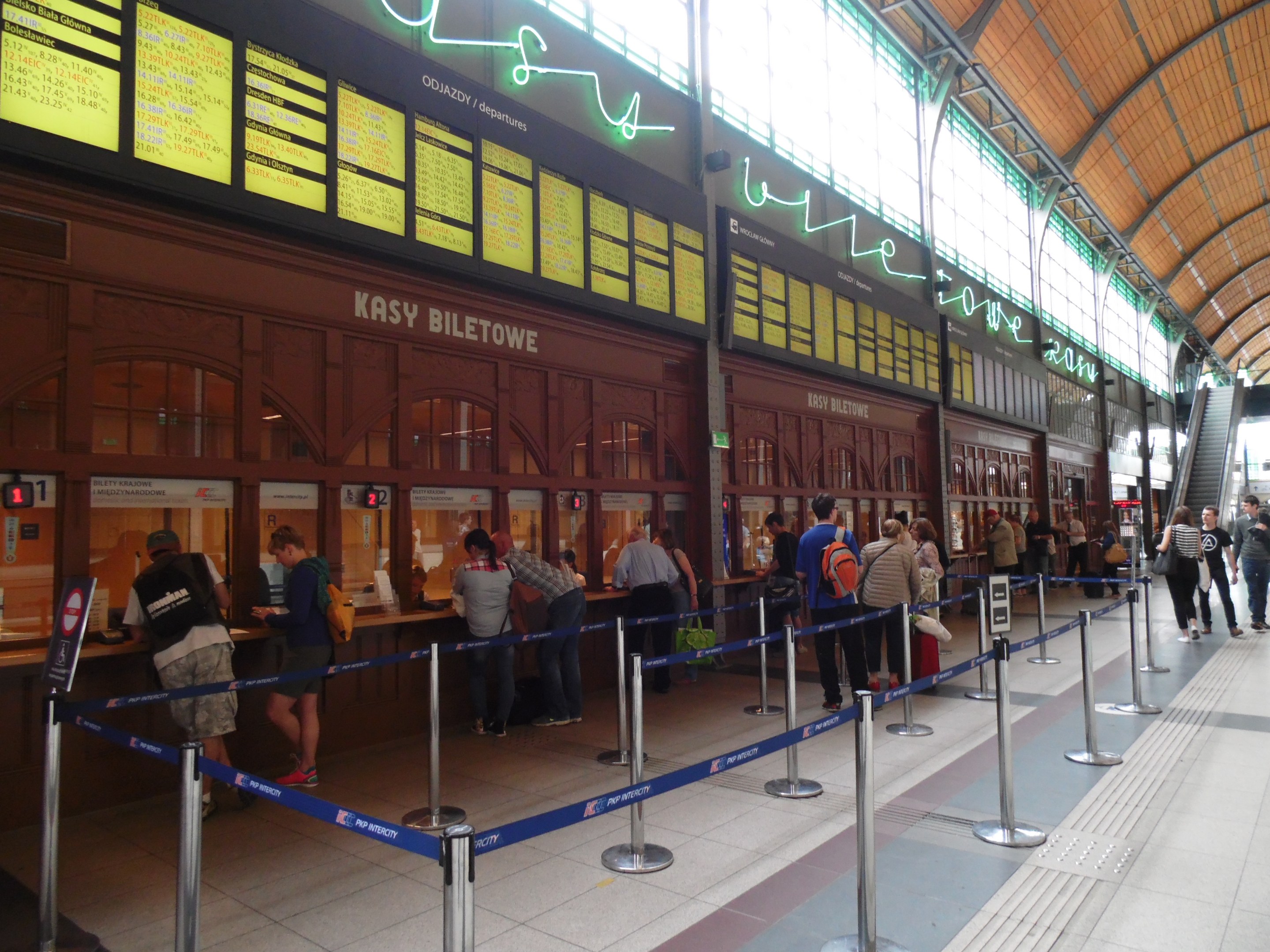 Menschen in einer Warteschlange an einem Bahnhofsschalter mit Stangen, Bändern, Informationsschildern, Deckenleuchten und Treppen mit Geländern im Hintergrund.