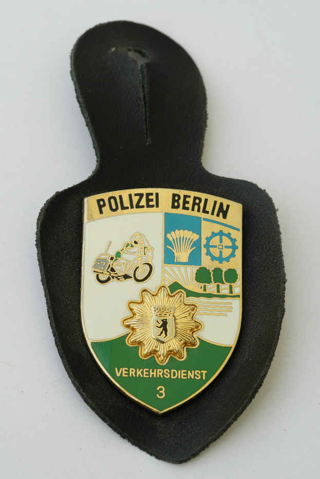 Ein schildförmiges Polizeibadges mit "Polizei Berlin" in fetter schwarzer Schrift auf einem blauen Hintergrund mit einem weißen Stern und Rahmen, das auf einer Oberfläche ruht.