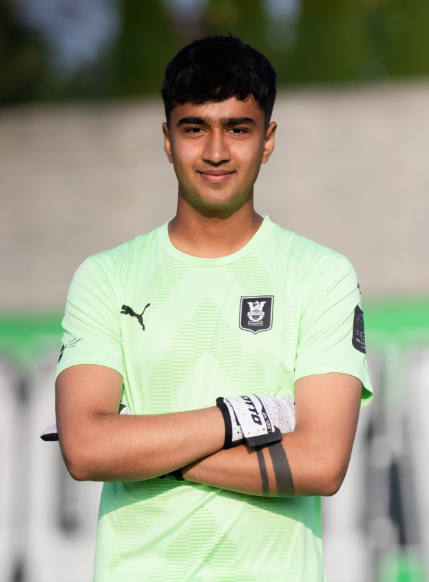Ein junger Mann in einem grünen Hemd steht lächelnd auf einem Fußballfeld mit verschränkten Armen, wahrscheinlich ein Torwart, mit einem unscharfen Hintergrund.