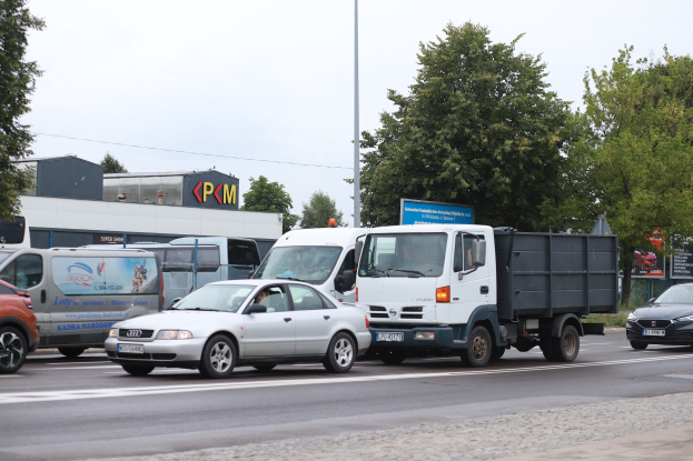 Eine Reihe von Fahrzeugen fährt eine von Bäumen gesäumte Straße mit Strommasten, Schildern, Gebäuden und einem klaren blauen Himmel im Hintergrund.