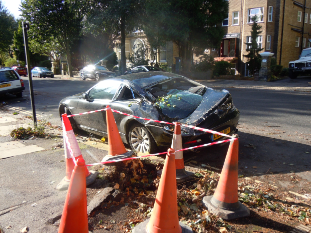 Ein besch├Ądigt aussehendes Auto am Stra├čenrand mit orangefarbenen Verkehrskegeln drumherum, Tannen und Geb├Ąude im Hintergrund unter einem klaren blauen Himmel.
