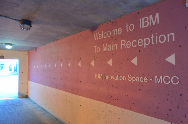 Eine Wand mit dem Text "Willkommen bei IBM", beleuchtet von Deckenlampen, mit einer Tür im Hintergrund, die zum IBM Innovation Space MCC führt.