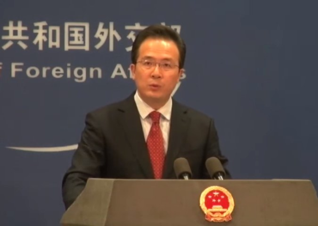 Ein Mann im Anzug und Krawatte steht an einem Rednerpult mit zwei Mikrofonen und hält eine Rede mit einem Logo auf dem Pult und einem Banner mit Text im Hintergrund.