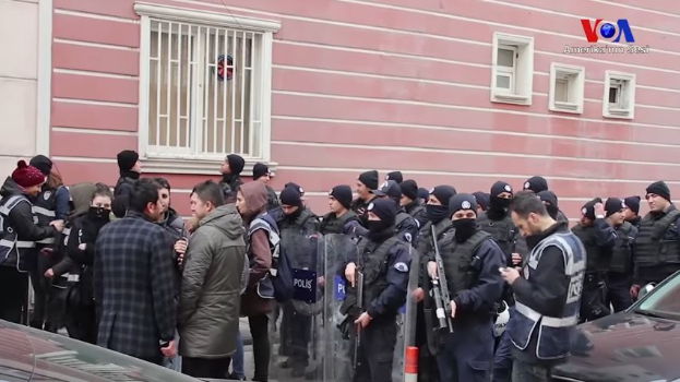 Eine Gruppe von Menschen, einige bewaffnet und mit Mützen, steht vor einem Gebäude mit Fenstern, versammelt, um gegen eine Regierungsentscheidung zu protestieren, mit Fahrzeugen und einem Verkehrskegel im Vordergrund und einem Wasserzeichen auf dem Bild.