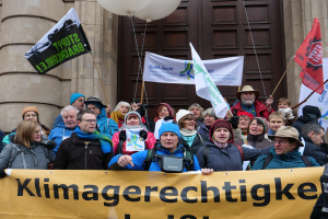 Eine Gruppe von Menschen vor einem Gebäude mit Säulen und einer Tür, die ein Transparent mit Text, Fahnen mit Stangen und Luftballons halten und an einem Klimastreik in Deutschland teilnehmen.