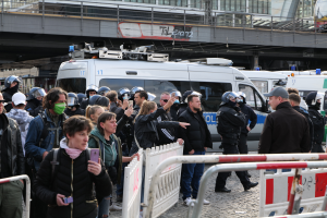 Eine Gruppe von Menschen steht vor Polizeiwagen, einige tragen Helme und halten Telefone, mit Barrieren im Vordergrund und einer Brücke mit Geländern und Gebäuden im Hintergrund in Berlin, Deutschland.