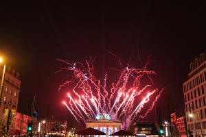 Eine belebte Stadtstraße in Berlin am Silvesterabend, voller Menschen, Fahrzeuge und Gebäude, beleuchtet von Feuerwerk und Gebäudelichtern, schafft eine festliche Atmosphäre.
