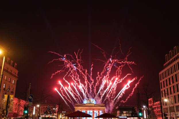 Eine belebte Stadtstraße in Berlin am Silvesterabend, voller Menschen, Fahrzeuge und Gebäude, beleuchtet von Feuerwerk und Gebäudelichtern, schafft eine festliche Atmosphäre.